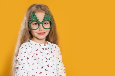 Girl wearing decorative eyeglasses in shape of Christmas trees on orange background, space for text