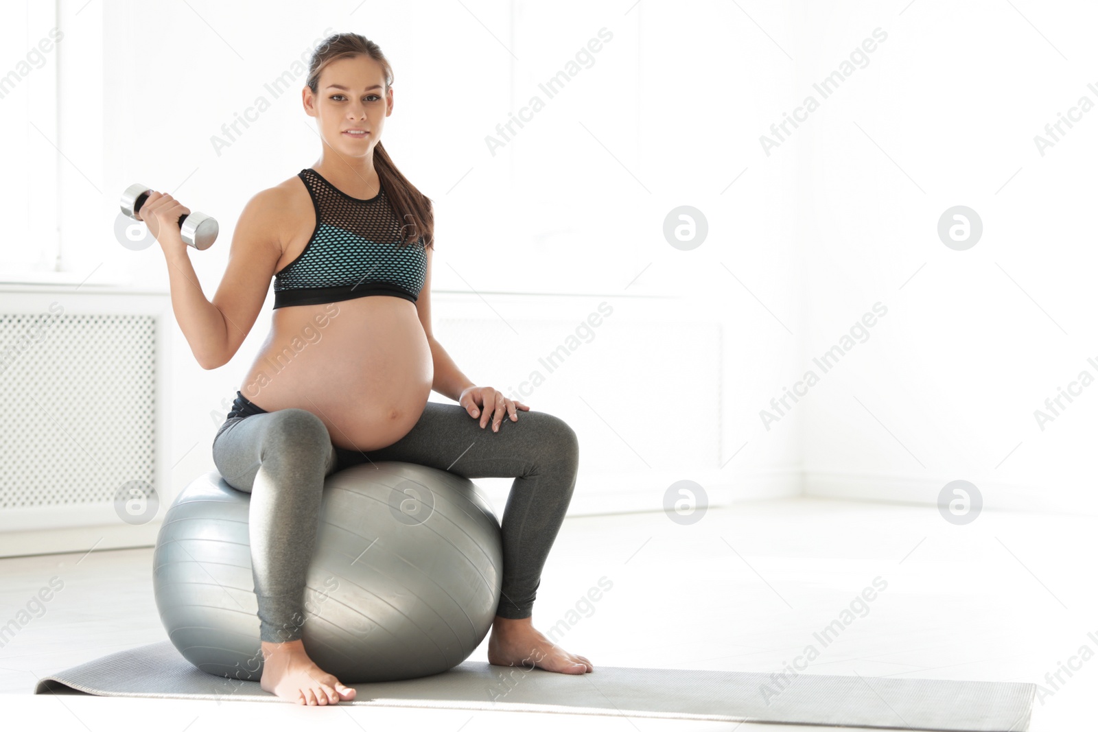 Photo of Young pregnant woman in fitness clothes lifting dumbbell at home. Space for text