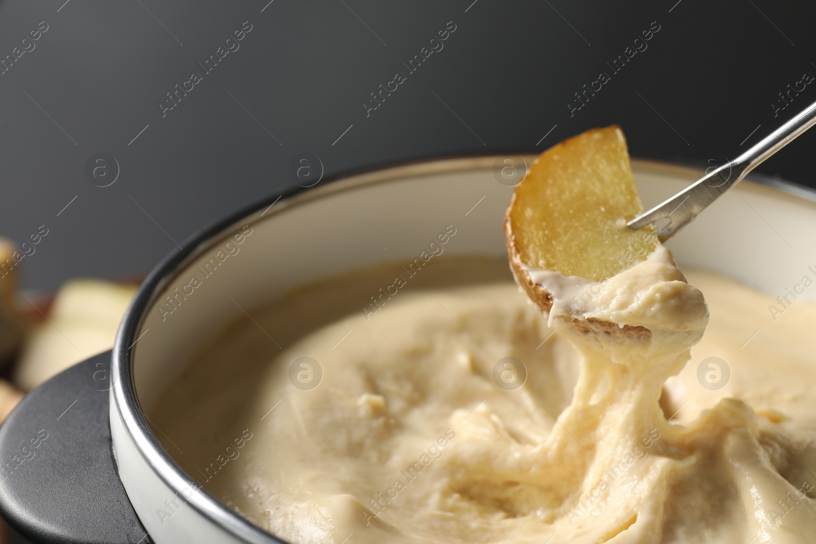 Photo of Dipping piece of potato into fondue pot with melted cheese on grey background, closeup