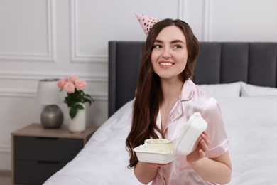 Photo of Beautiful young woman in party hat holding her Birthday cake on bed at home