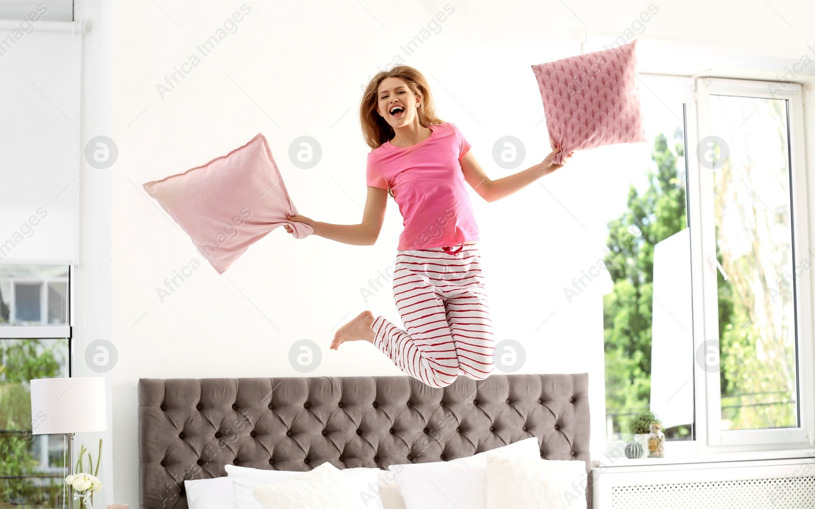 Photo of Young woman with pillows jumping on bed at home