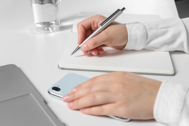 Photo of Woman with smartphone writing in notebook at white table, closeup
