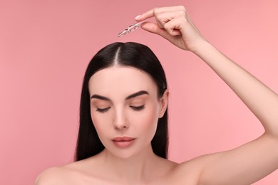 Beautiful young woman using ampoule for hair treatment on pink background