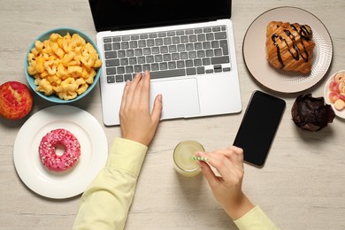 Photo of Bad eating habits. Woman with glass of drink and different snacks using laptop at wooden table, top view