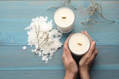 Photo of Woman with burning soy candles at light blue wooden table, top view