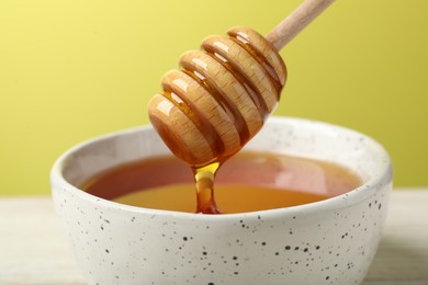 Photo of Pouring tasty honey from dipper into bowl at white table against yellow background, closeup