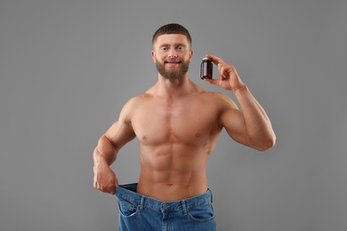 Photo of Athletic young man with bottle of supplements wearing big jeans on grey background. Weight loss