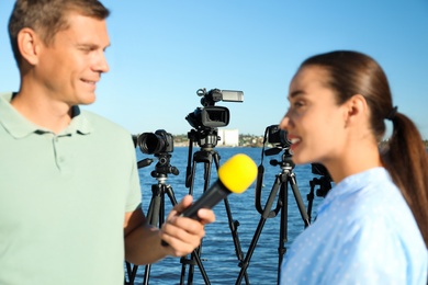 Photo of Journalist interviewing young woman near river, focus on video cameras