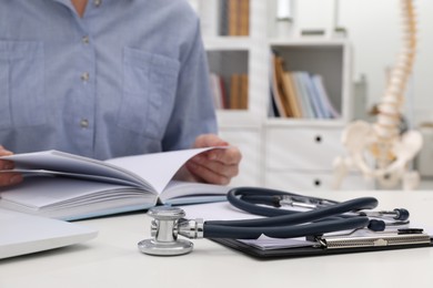 Photo of Woman with book, stethoscope and clipboard at white table indoors, closeup. Medical education