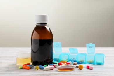 Photo of Bottle of syrup, measuring cup, dosing spoon and pills on white wooden table against light grey background. Cold medicine