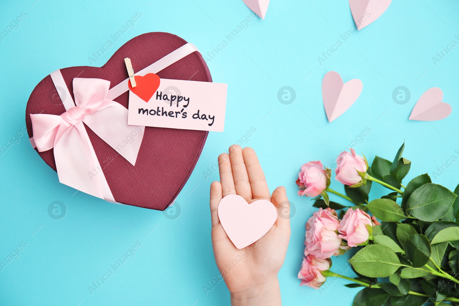 Photo of Child holding small paper heart near gift box and roses for Mother's day on color background