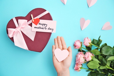 Child holding small paper heart near gift box and roses for Mother's day on color background