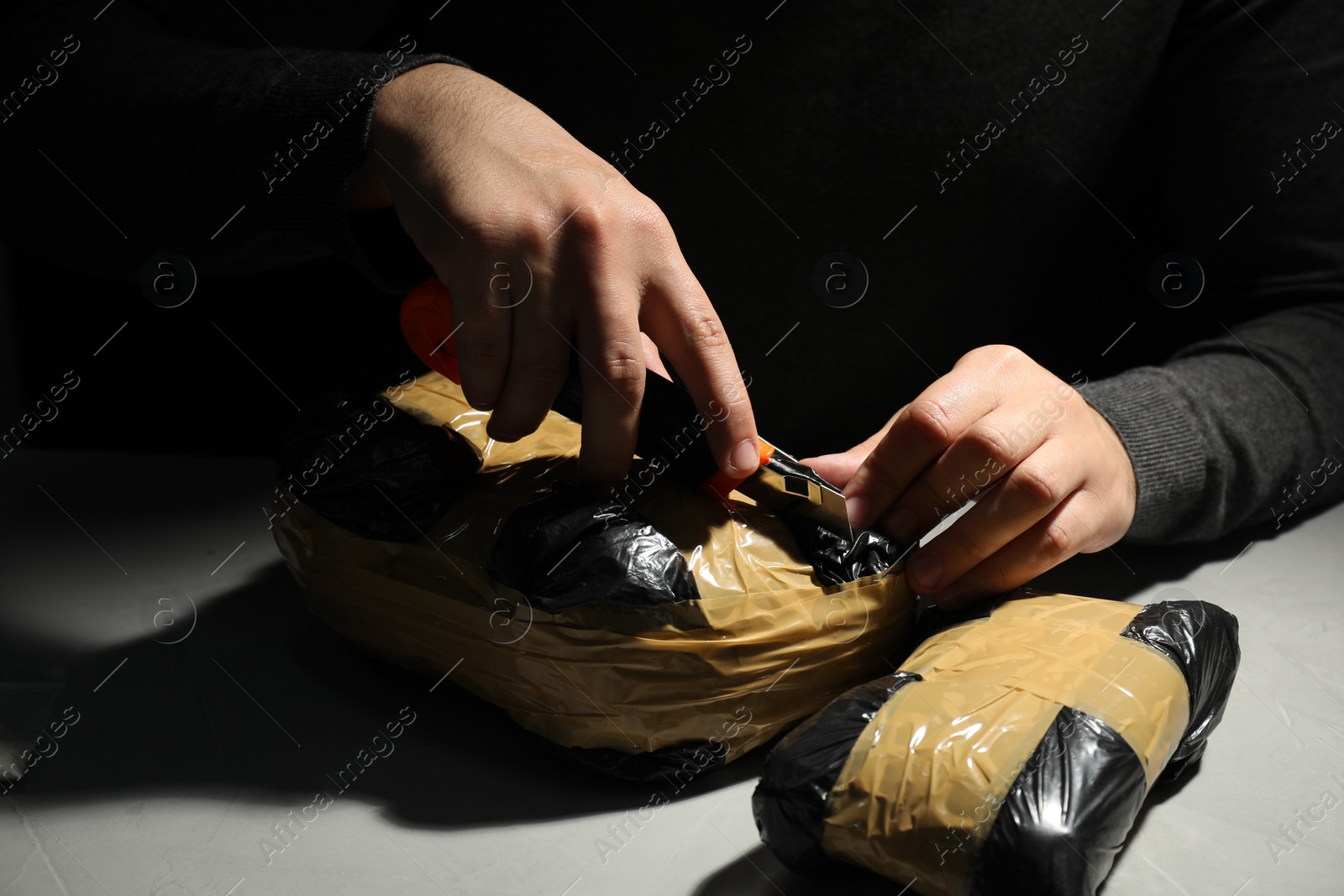 Photo of Smuggling and drug trafficking. Man opening package of narcotics with box cutter in darkness, closeup