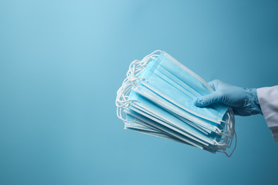 Photo of Doctor in latex gloves holding disposable face masks on light blue background, closeup with space for text. Protective measures during coronavirus quarantine