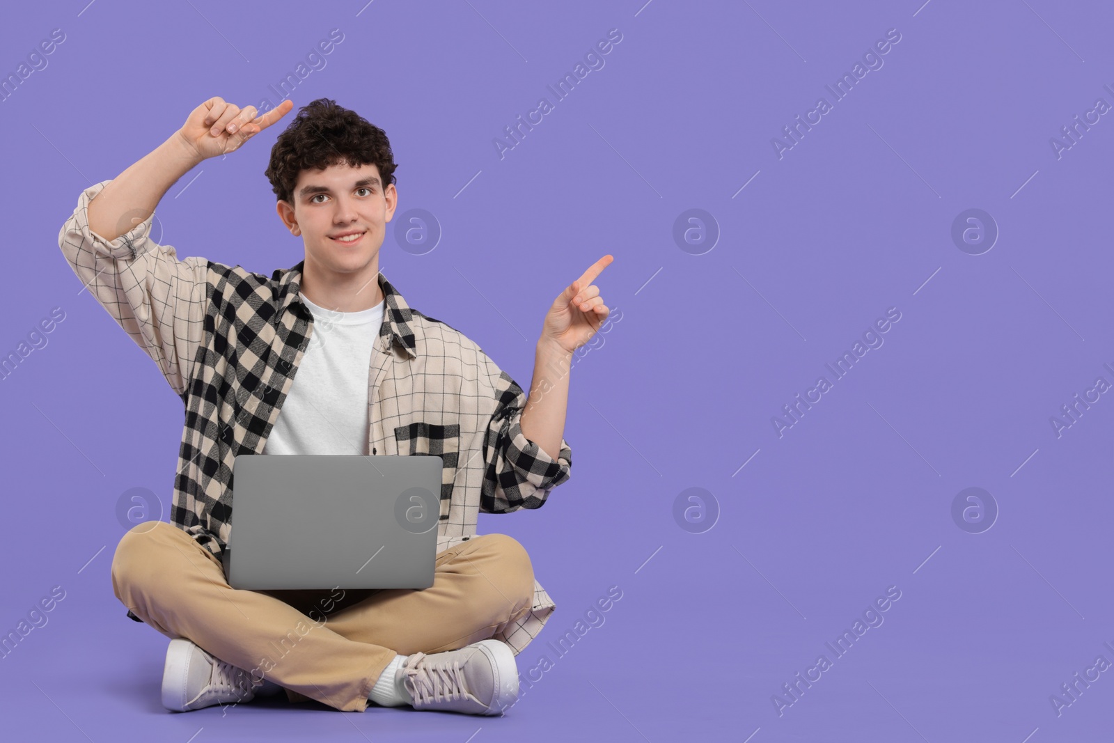 Photo of Portrait of student with laptop sitting and pointing on purple background. Space for text