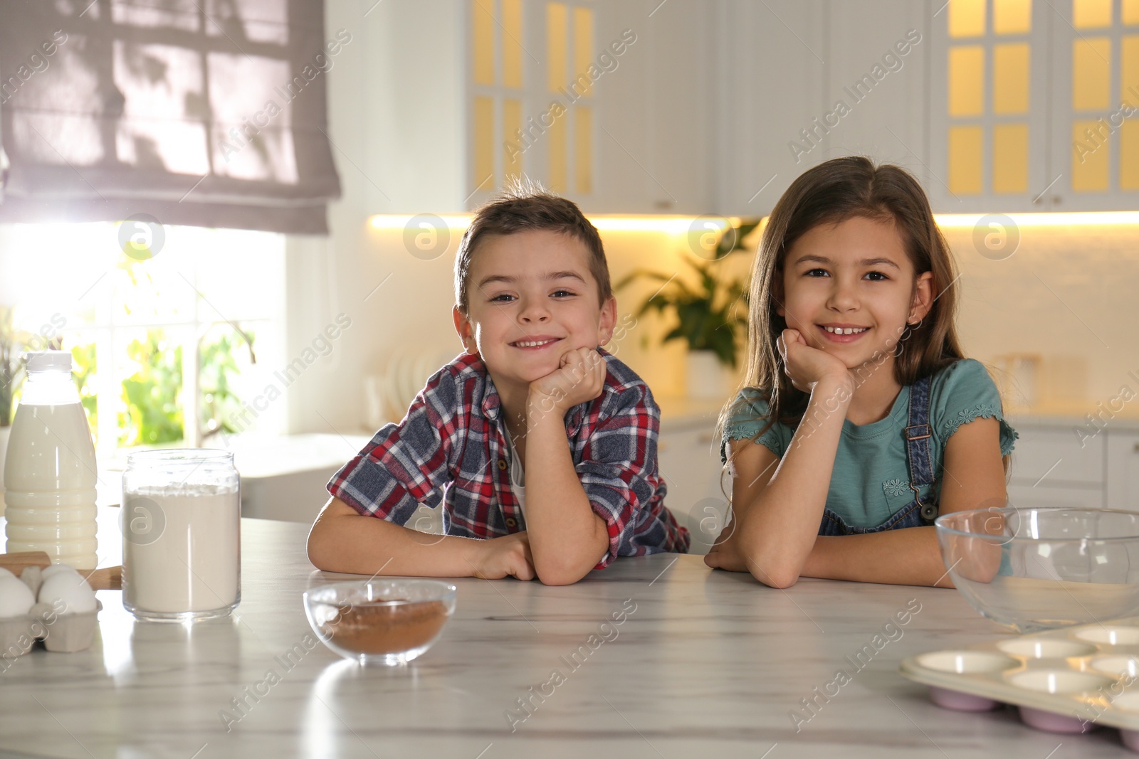 Photo of Cute little children at table with cooking ingredients in kitchen