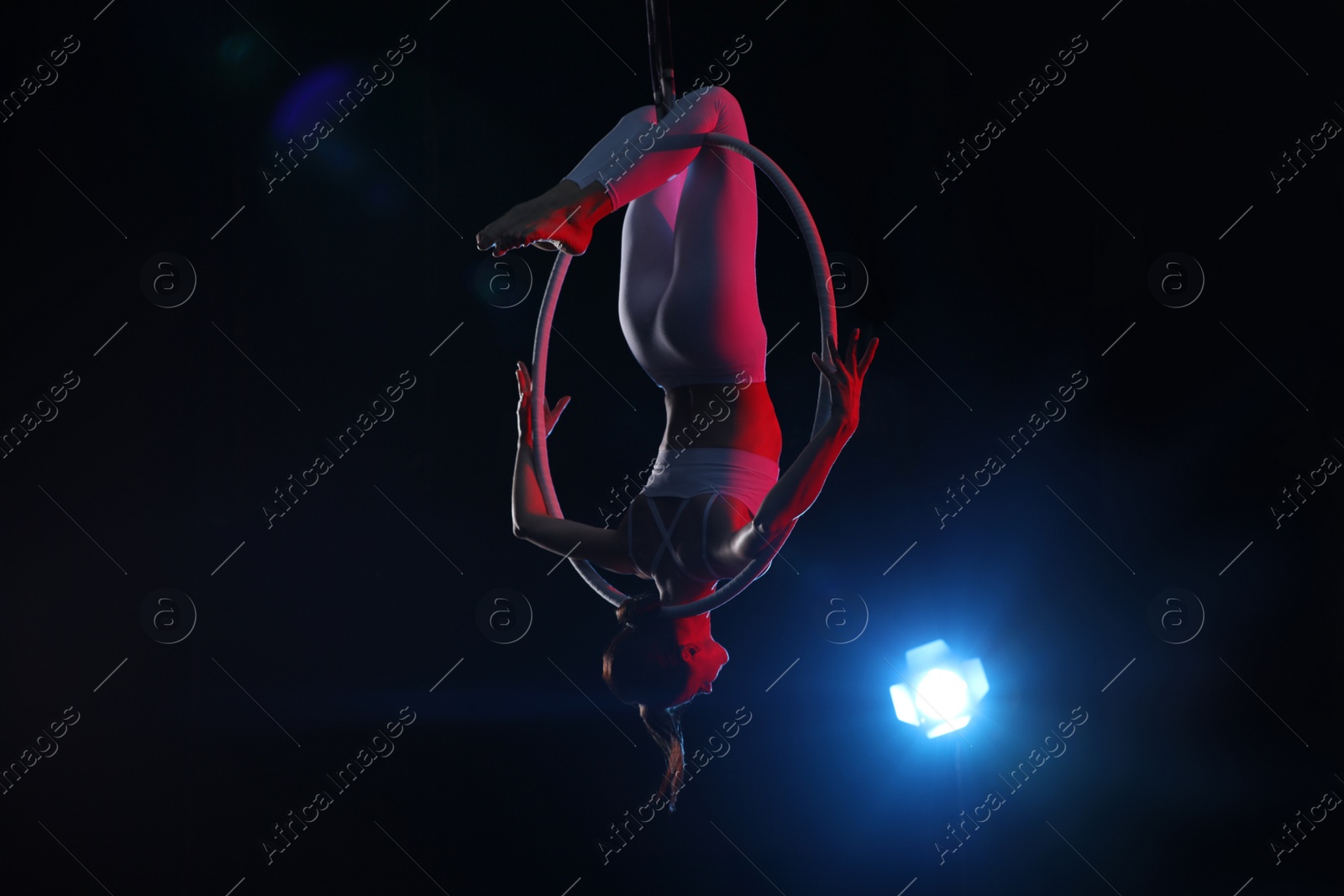 Photo of Young woman performing acrobatic element on aerial ring against dark background