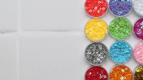 Many different colorful sequins in containers on white tiled background, top view. Space for text