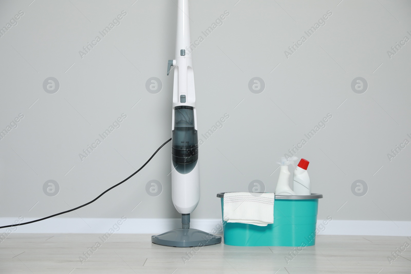Photo of Modern steam mop and bucket with different cleaning supplies on floor near grey wall