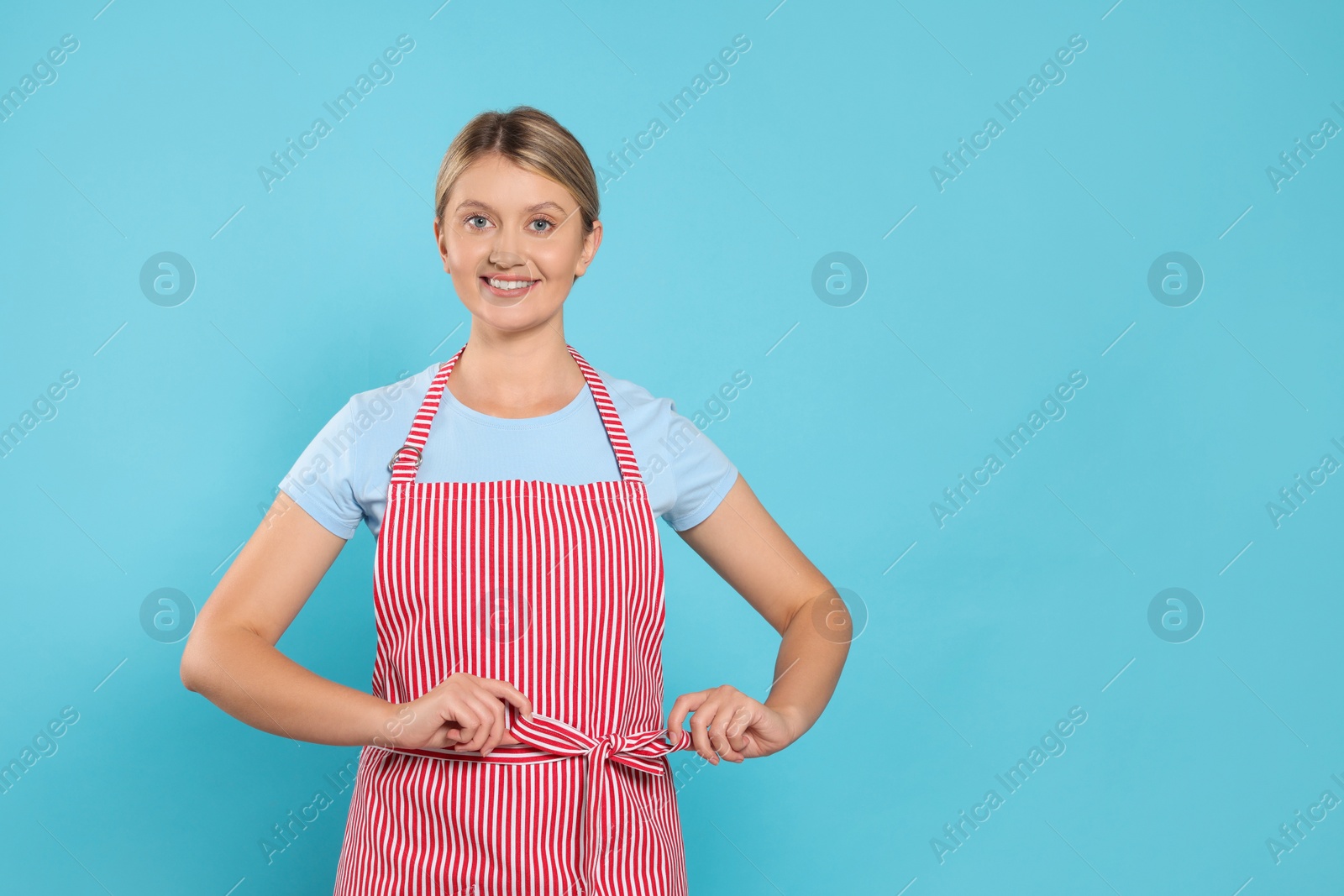 Photo of Beautiful young woman in clean striped apron on light blue background. Space for text