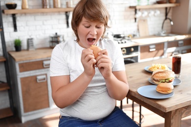Photo of Emotional overweight boy at table with fast food in kitchen