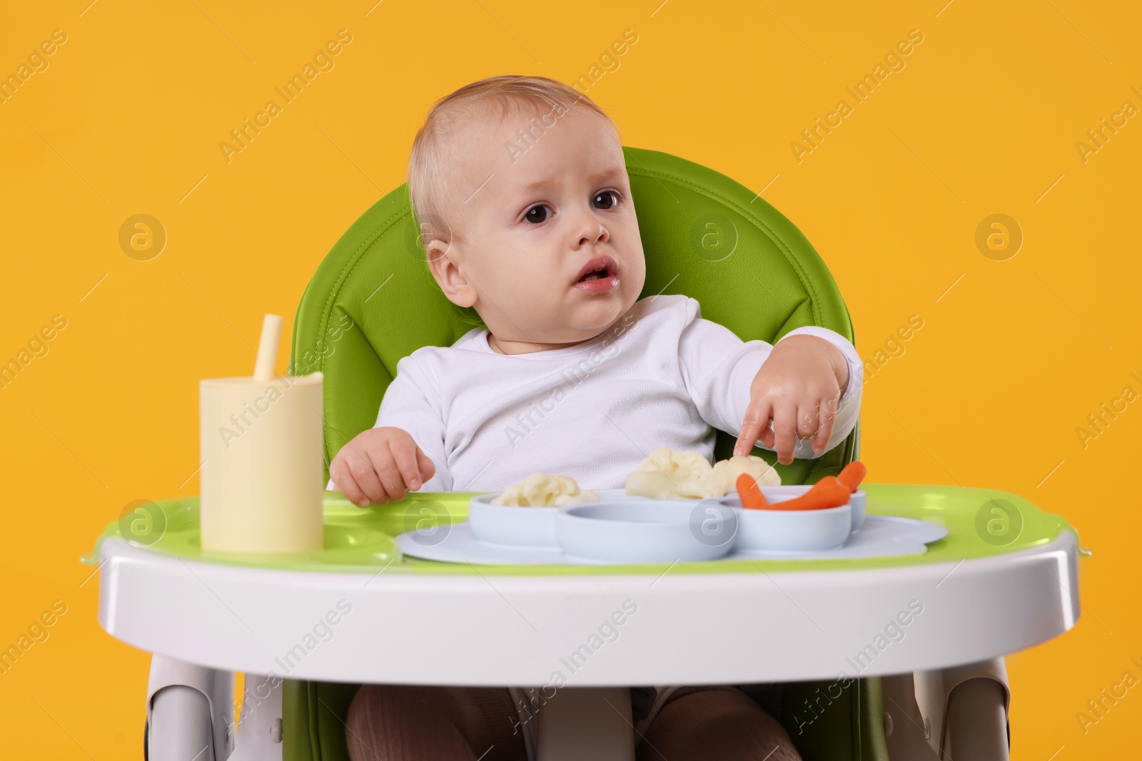 Photo of Cute little baby with healthy food in high chair on orange background