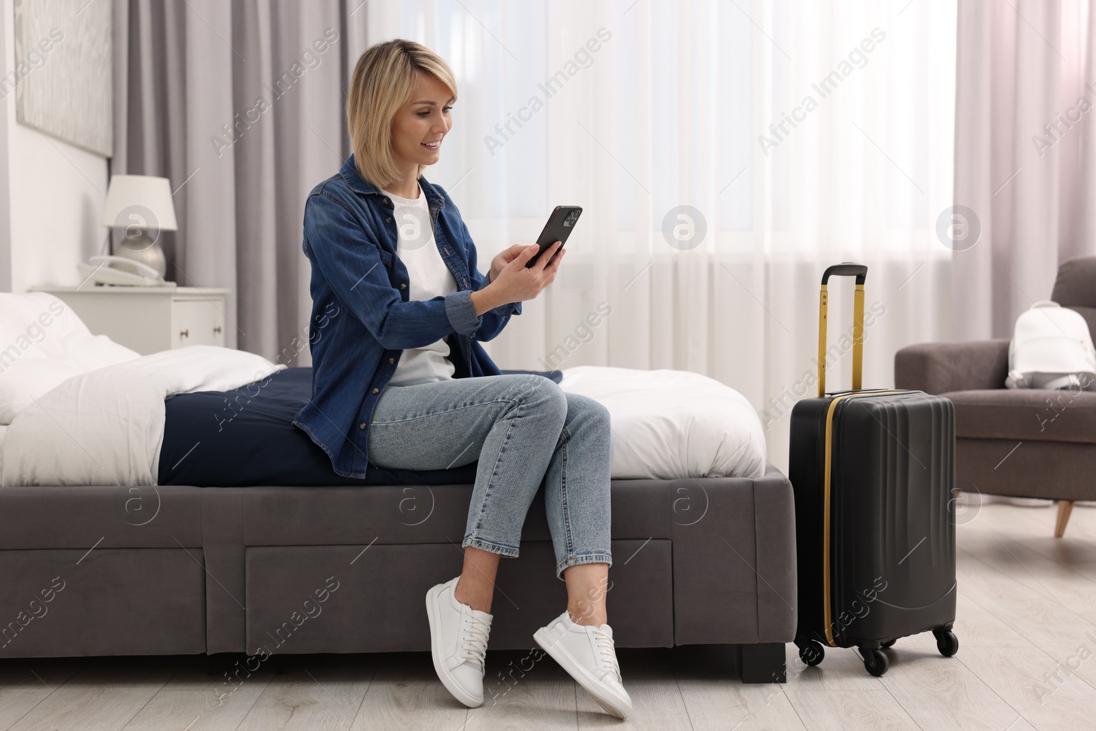 Photo of Smiling guest with smartphone and suitcase relaxing on bed in stylish hotel room