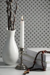 Photo of Books, burning candle, rosary beads and vase of willow branches on white table, closeup