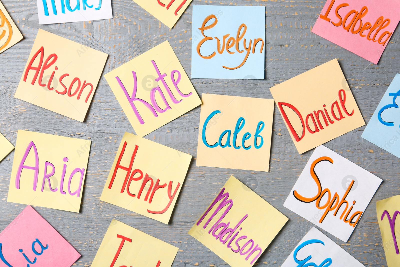 Photo of Colorful paper sheets with written different baby names on grey wooden table, flat lay