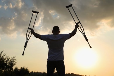 Man raising hands with underarm crutches up to sky outdoors at sunset, back view. Healing miracle