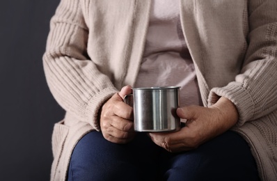 Photo of Poor elderly woman holding metal mug on dark background, closeup