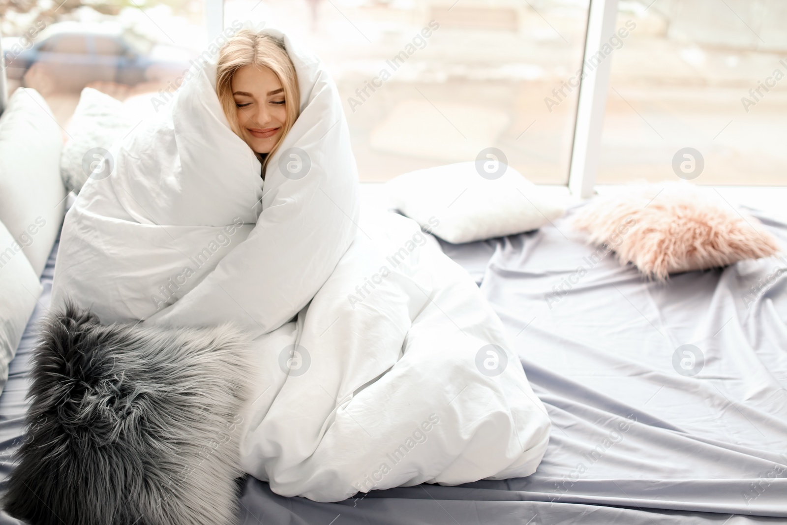 Photo of Beautiful young woman wrapped in soft blanket sitting near window at home. Winter atmosphere