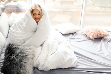 Photo of Beautiful young woman wrapped in soft blanket sitting near window at home. Winter atmosphere