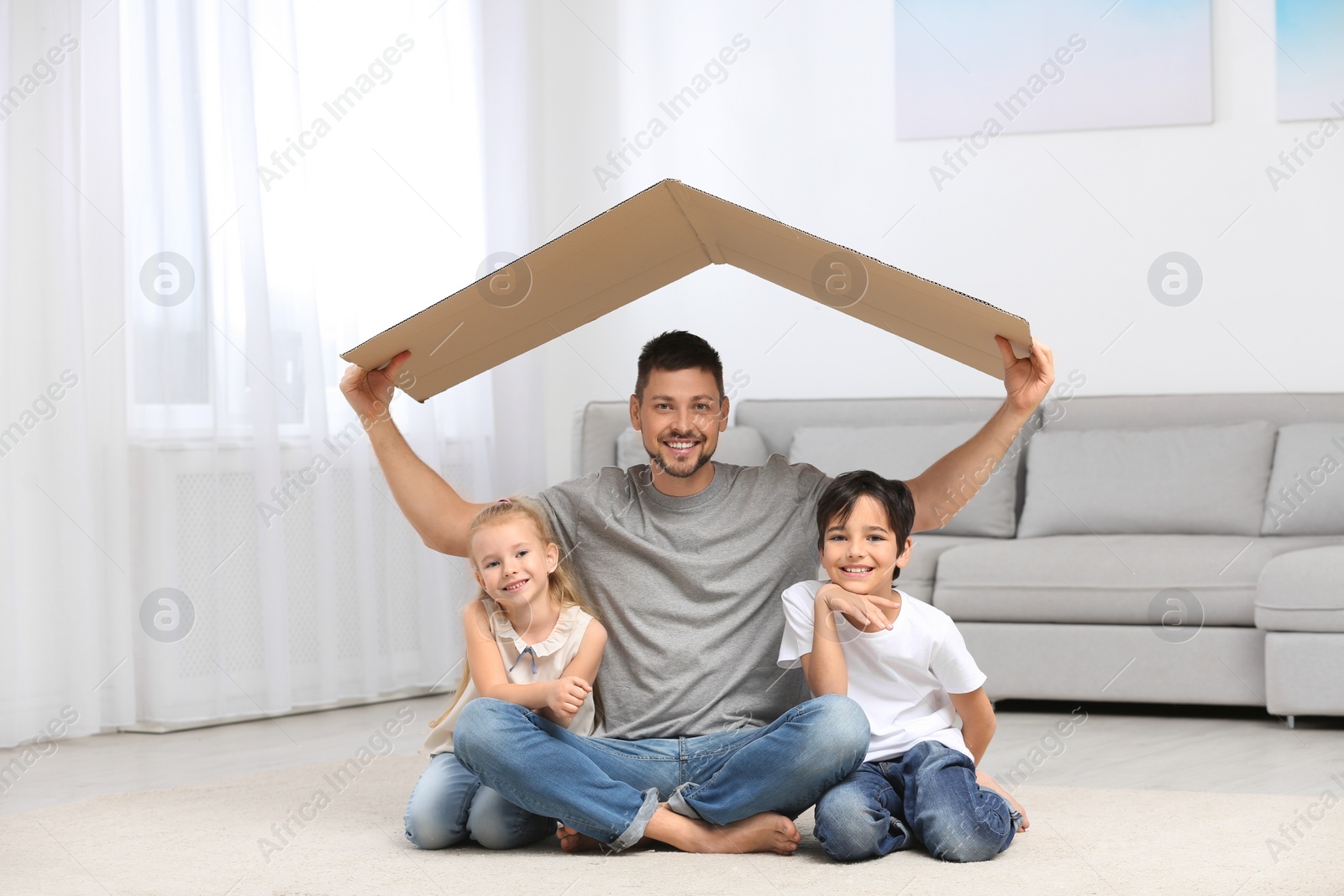 Photo of Happy father and his children sitting under cardboard roof at home. Insurance concept
