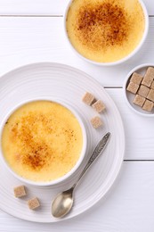 Photo of Delicious creme brulee in bowls, sugar cubes and spoon on white wooden table, flat lay