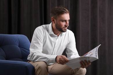 Handsome man reading magazine in armchair against dark background