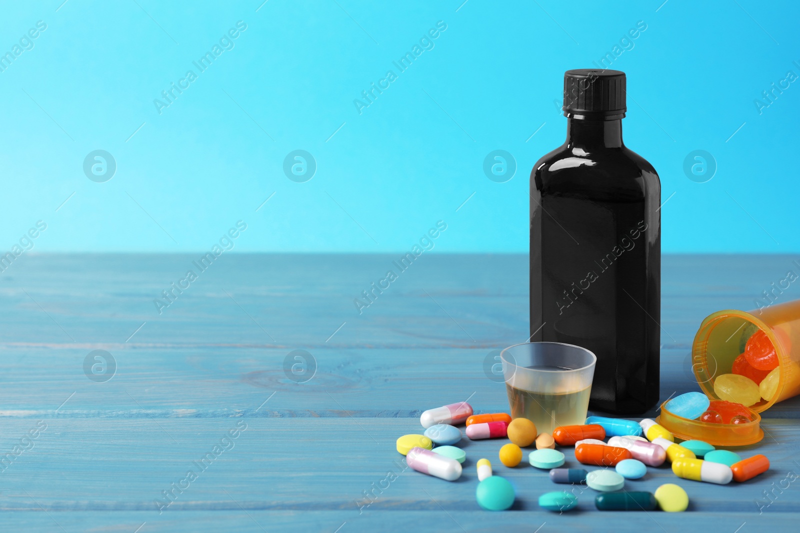 Photo of Bottle of syrup, measuring cup and pills on light blue table, space for text. Cold medicine