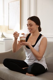 Woman eating tasty granola with fresh berries and yogurt on sofa at home