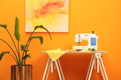 Photo of Modern sewing machine with cloth and craft accessories on wooden table near orange wall in room