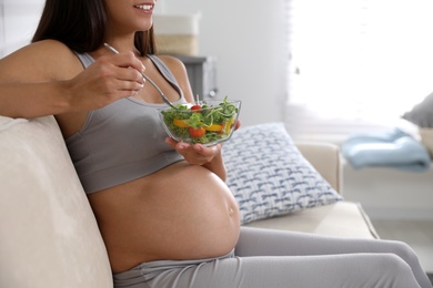 Young pregnant woman with bowl of vegetable salad in living room, closeup. Taking care of baby health