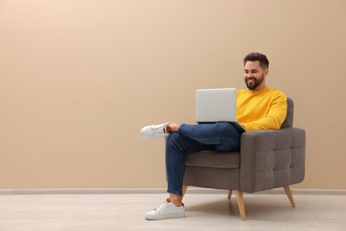 Photo of Handsome man with laptop sitting in armchair near beige wall indoors, space for text