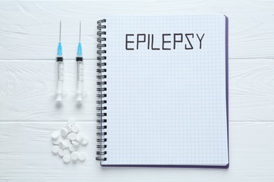 Photo of Notebook with word Epilepsy, pills and syringes on white wooden table, flat lay