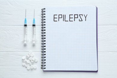 Notebook with word Epilepsy, pills and syringes on white wooden table, flat lay