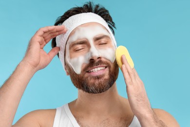 Man with headband washing his face using sponge on light blue background