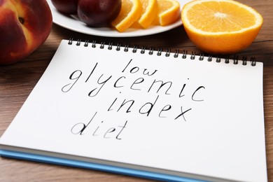 Photo of Notebook with words Low Glycemic Index Diet and fresh fruits on wooden table, closeup