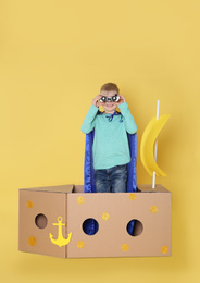 Little child in cape playing with ship made of cardboard box on yellow background