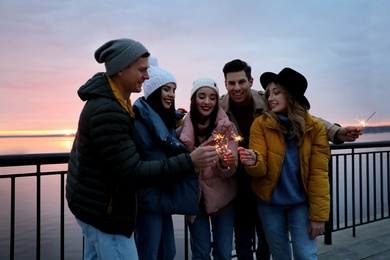 Photo of People in warm clothes holding burning sparklers near river