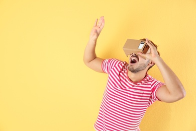 Young man using cardboard virtual reality headset on color background. Space for text