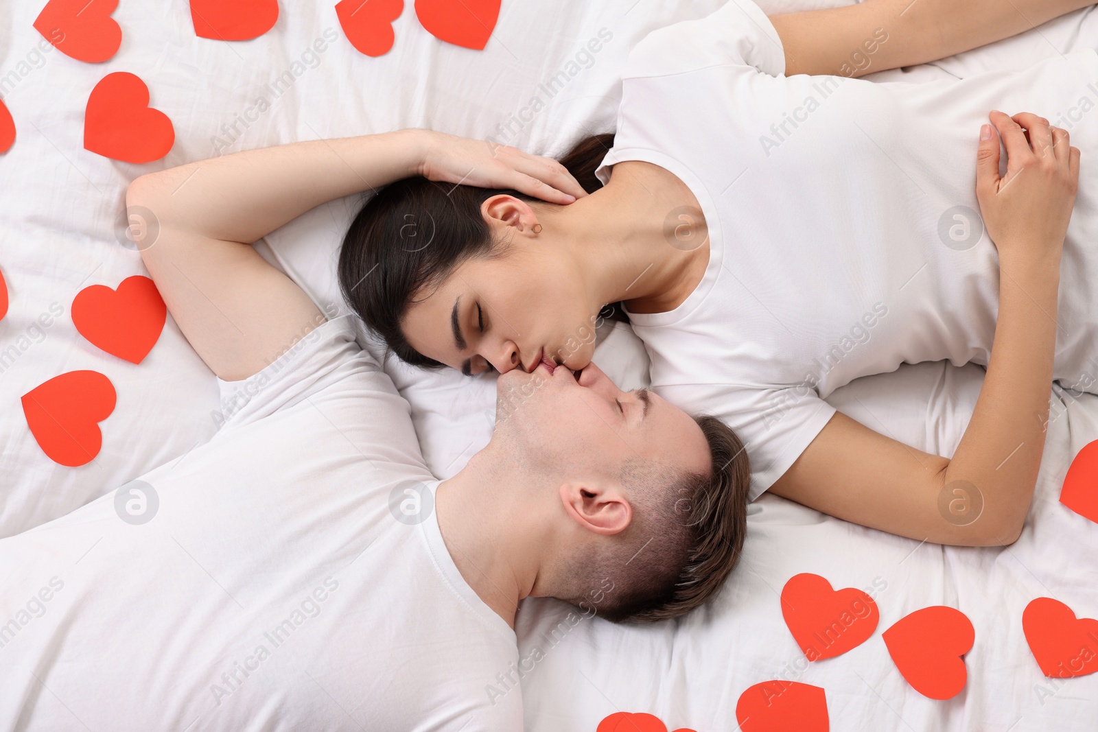 Photo of Lovely couple kissing on bed with many decorative hearts, top view. Valentine's day celebration