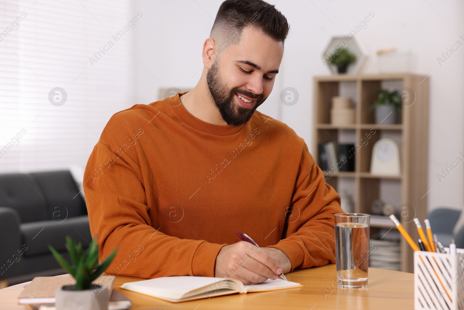 Photo of Young man writing in notebook at wooden table indoors